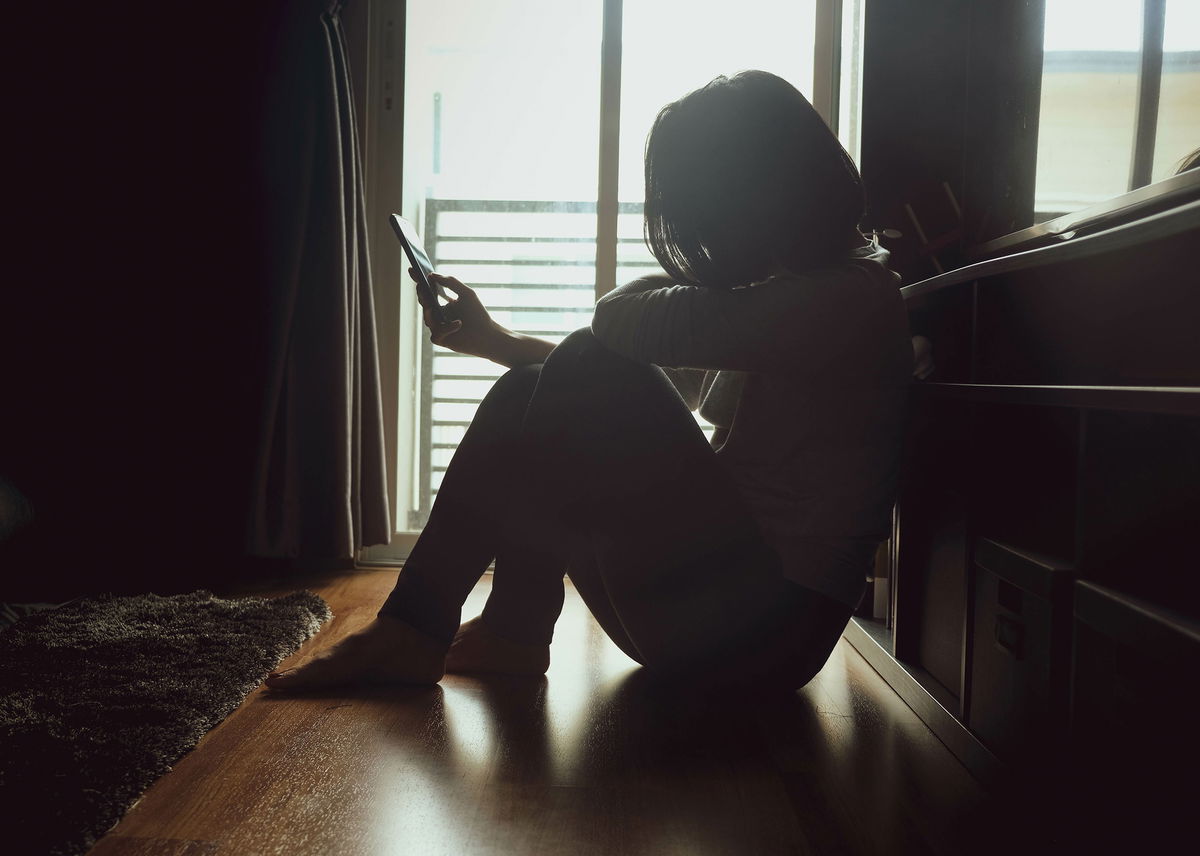 Asian women with short and black hair in casual wear, sitting on a wooden floor side the window with some light in dark tone room and holding a mobile phone. Maybe she's cheating something to somebody