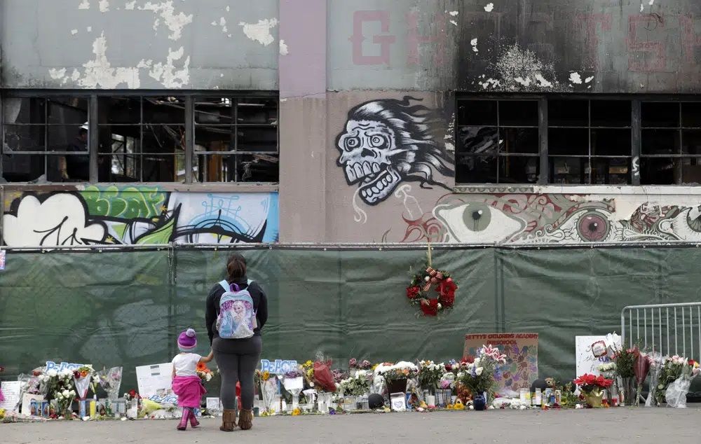 FILE - Flowers, pictures, signs and candles, are placed at the scene of a warehouse fire which killed dozens in Oakland, Calif. Dec. 13, 2016. The converted artists' warehouse that was acquired by The Unity Council, a nonprofit community development organization, was quietly razed in May 2023, in preparation for possible development into badly needed low-income housing