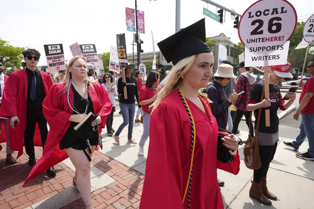 People dressed in commencement gowns, front, walk past protesters supporting the Hollywood writers' strike outside an entrance to Boston University commencement ceremonies, Sunday, May 21, 2023, in Boston. David Zaslav, president and CEO of Warner Bros. Discovery, delivered an address during the ceremonies Sunday.