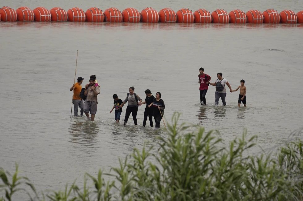 Migrants who crossed the Rio Grande from Mexico walk past large buoys being deployed as a border barrier on the river in Eagle Pass, Texas on Wednesday July 12, 2023. The floating barrier is being deployed in an effort to block migrants from entering Texas from Mexico